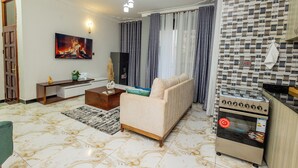 A living room with a sofa, a coffee table, a TV, and a kitchen area with a stove. - The Nest Kampala (Kampala)