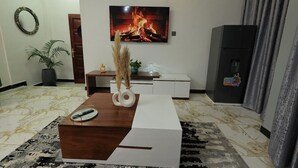 A modern living room with a flat-screen TV mounted on the wall, a wooden coffee table, a patterned rug, and a minimalist console table. - The Nest Kampala (Kampala)