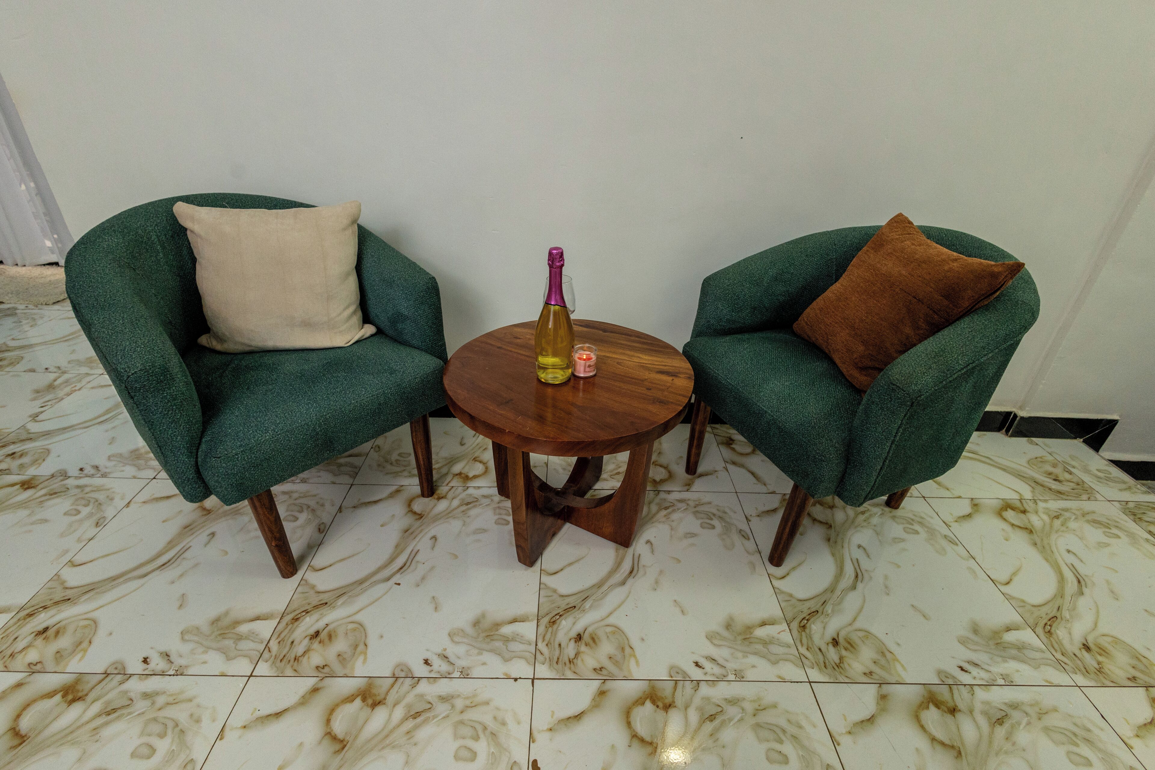 Two green armchairs with different colored cushions, a small wooden table with a bottle and a candle, and a marble floor.