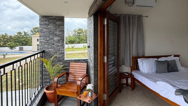 A balcony with a wooden chair, a small table with a vase and a plant, and a bed with pillows. - Breezy villa (Shrirangapattana)