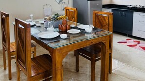 A dining area with a glass table and wooden chairs, a kitchen with dark cabinets and a stainless steel refrigerator, and a window with blinds. - Breezy villa (Shrirangapattana)
