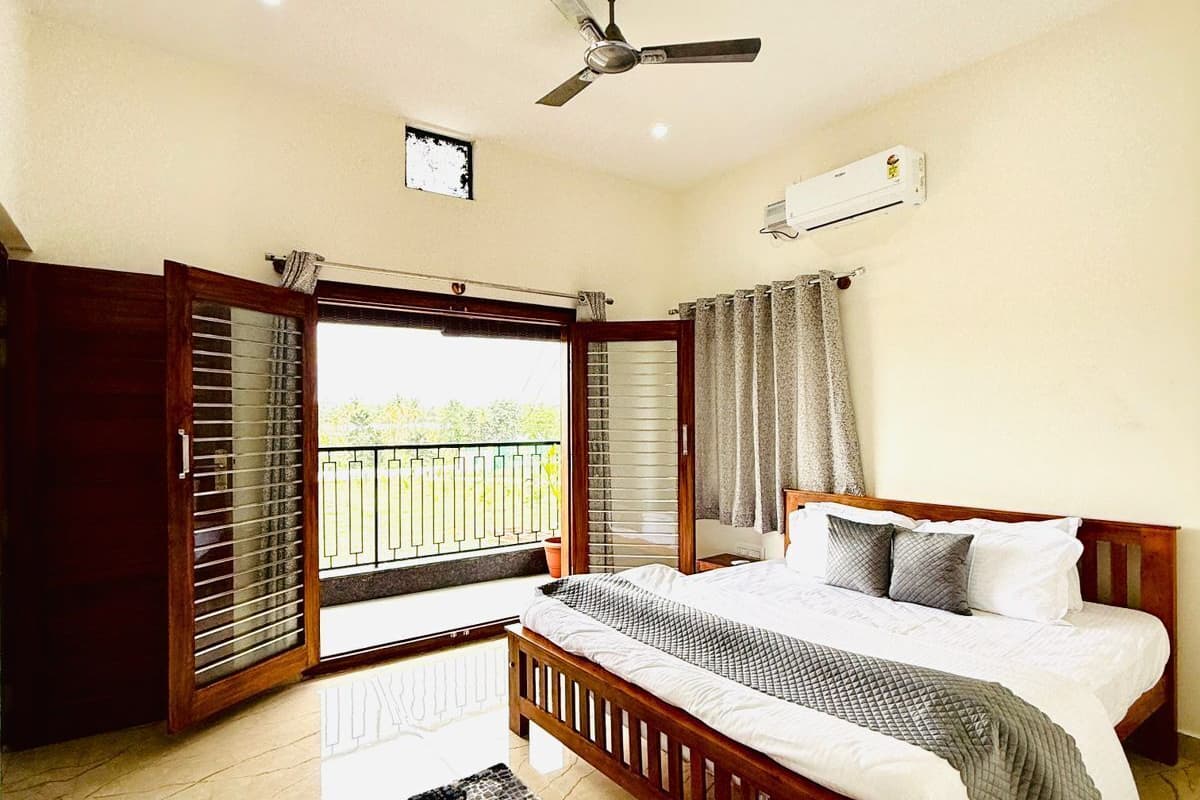 A bedroom with a bed, a ceiling fan, an air conditioning unit, a balcony with a view, and a patterned rug.