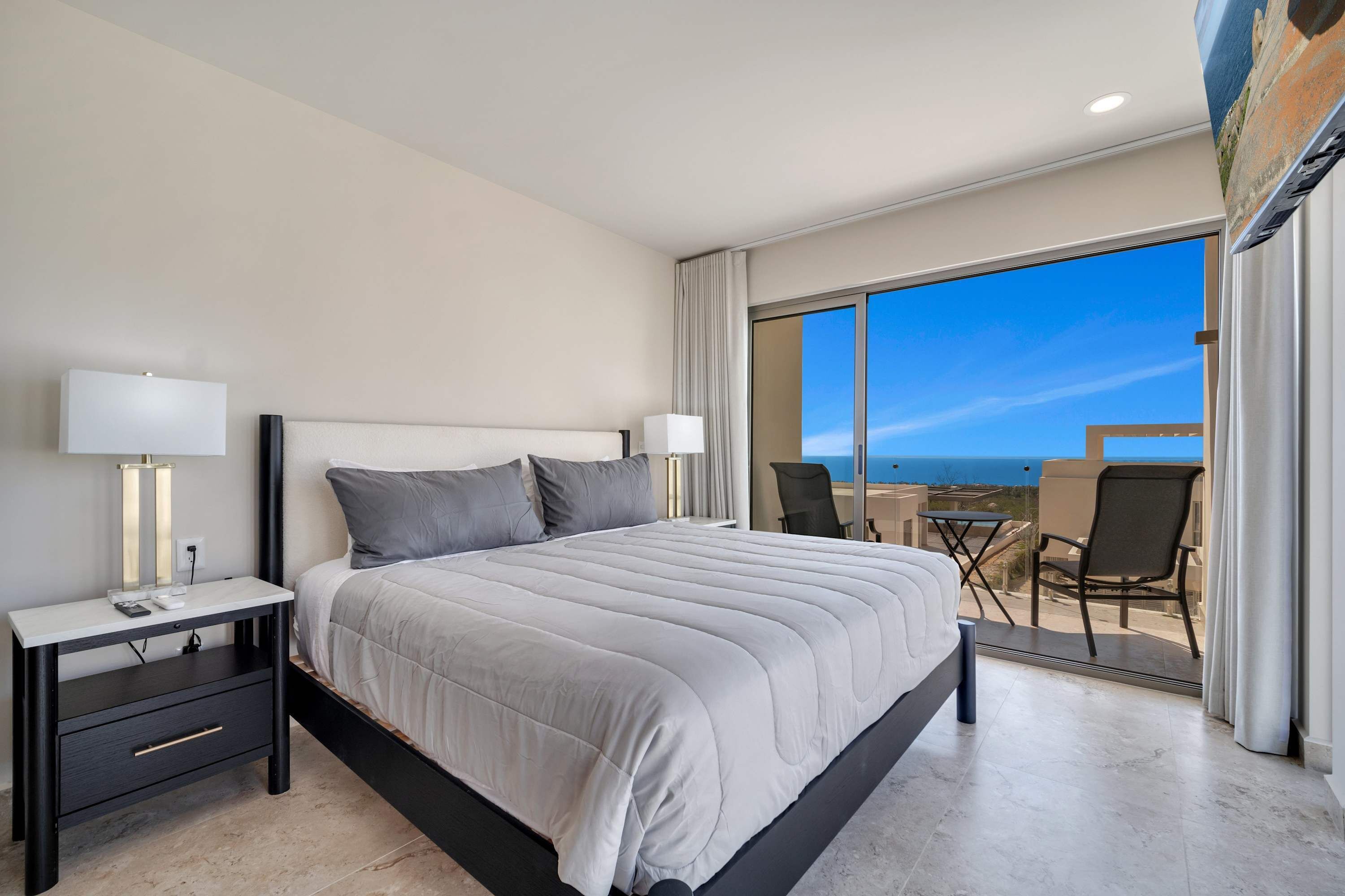 A modern hotel room with a large bed, two bedside tables with lamps, a balcony with a table and chairs, and a view of the sea.
