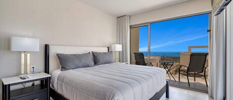 A modern hotel room with a large bed, two bedside tables with lamps, a balcony with a table and chairs, and a view of the sea.
