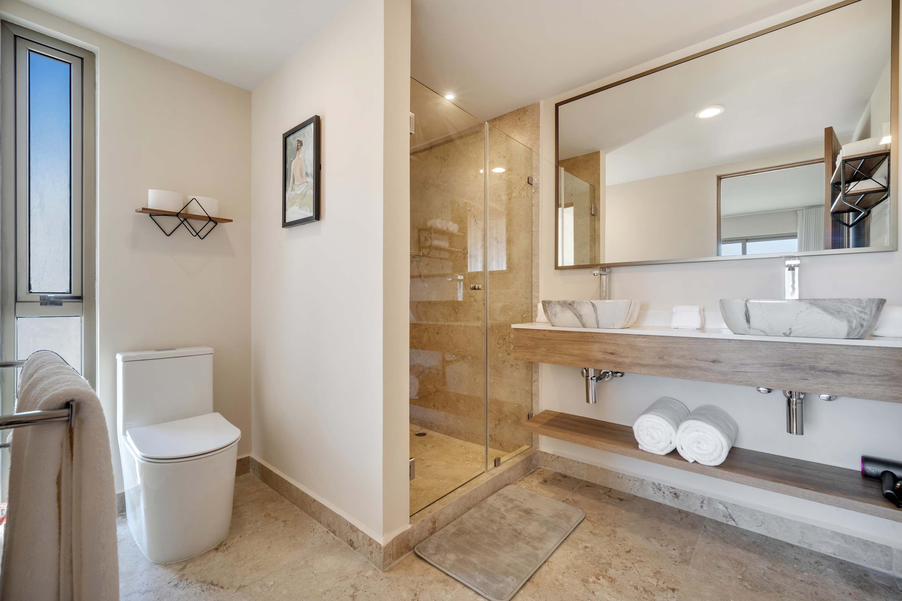 A modern bathroom with a glass shower enclosure, a white toilet, a large mirror, and a wooden vanity with marble accents.