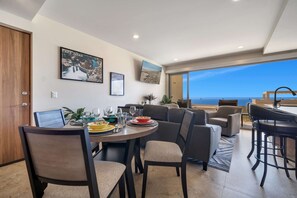 A dining area with a table set for four, a bar area with stools, and a large window offering a view of the ocean. - Stunning Brand New Condo - Pool & Ocean Views (Cabos San Lucas)
