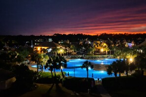 A resort with a large swimming pool, palm trees, and illuminated buildings at dusk.