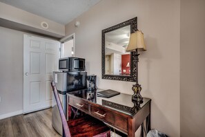 A room with a wooden desk, a mirror, a lamp, a microwave, and a refrigerator.