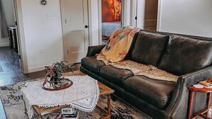 Sala de estar com sofá de couro marrom, mesinha de centro e tapete estampado. Uma porta dá acesso a outro quarto com cama e aquecedor na parede.