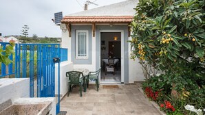 Una casa con techo de tejas, un portón azul, un pequeño patio con mesa y sillas, y un jardín con árboles frutales y flores.