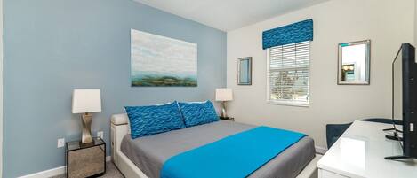 A bedroom with a bed, two bedside lamps, a desk with a computer, a window with blinds, and a painting on the wall.