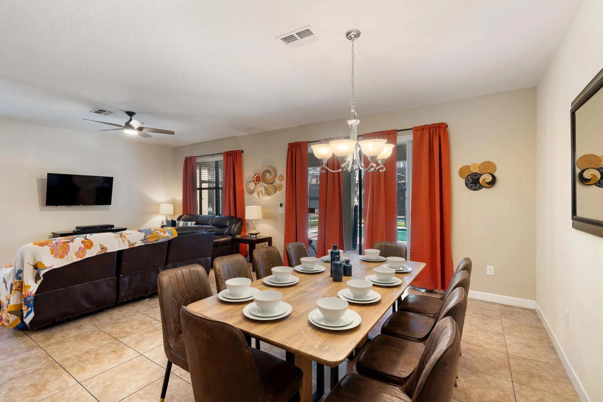 A dining area with a long table set for six, surrounded by brown leather chairs. A television is mounted on the wall to the left, and a ceiling fan is visible above.