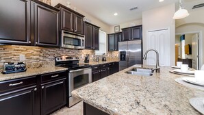 A modern kitchen with dark cabinetry, granite countertops, stainless steel appliances, and a central island with a sink. - BUC2979 (Kissimmee)
