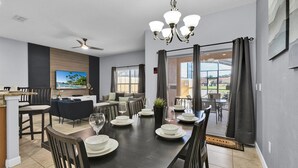 Un comedor moderno con una mesa de madera oscura dispuesta para cuatro personas, una araña y un televisor montado en la pared.
