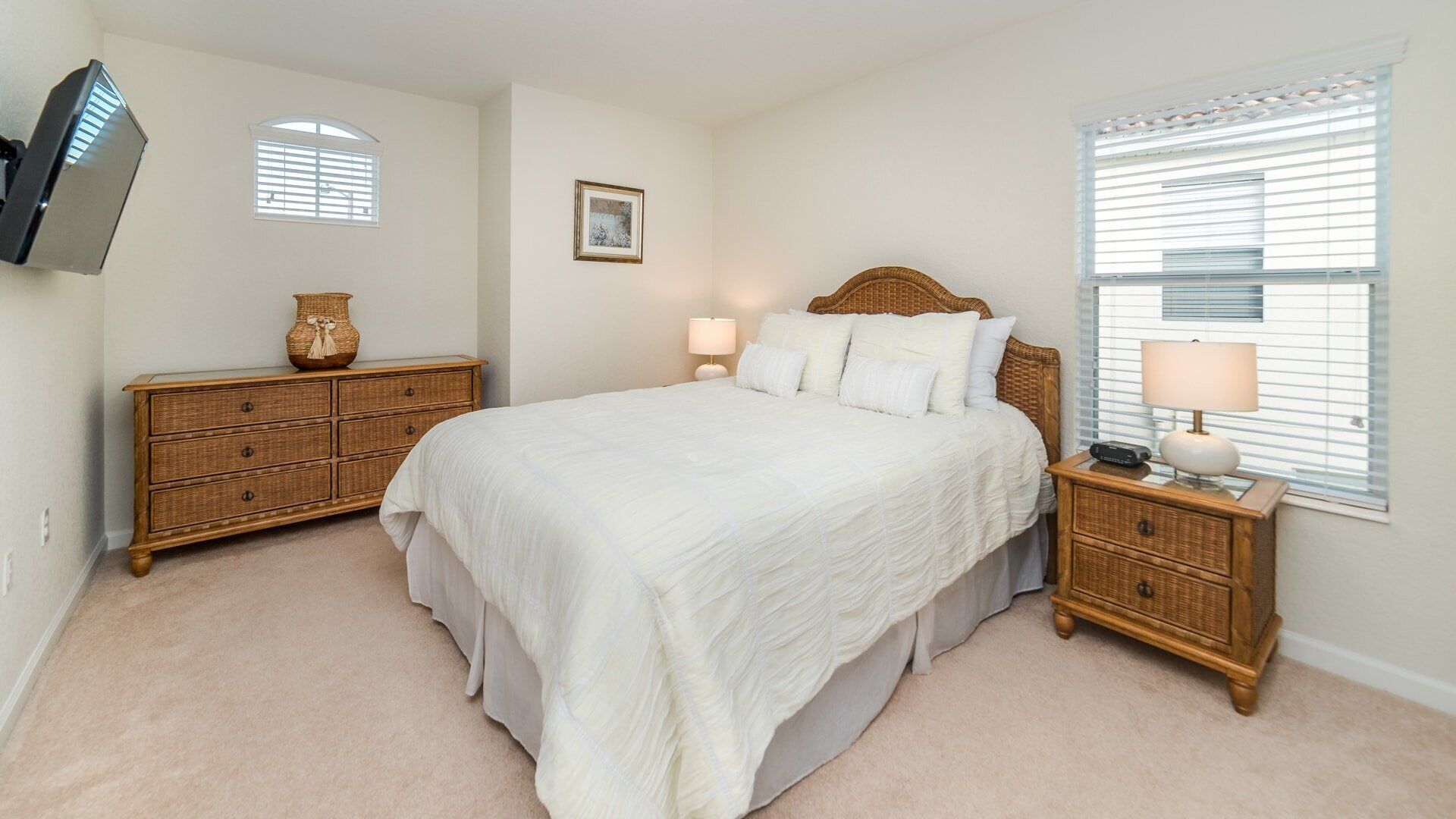 A bedroom with a large bed, two nightstands, a television mounted on the wall, and a window with blinds.