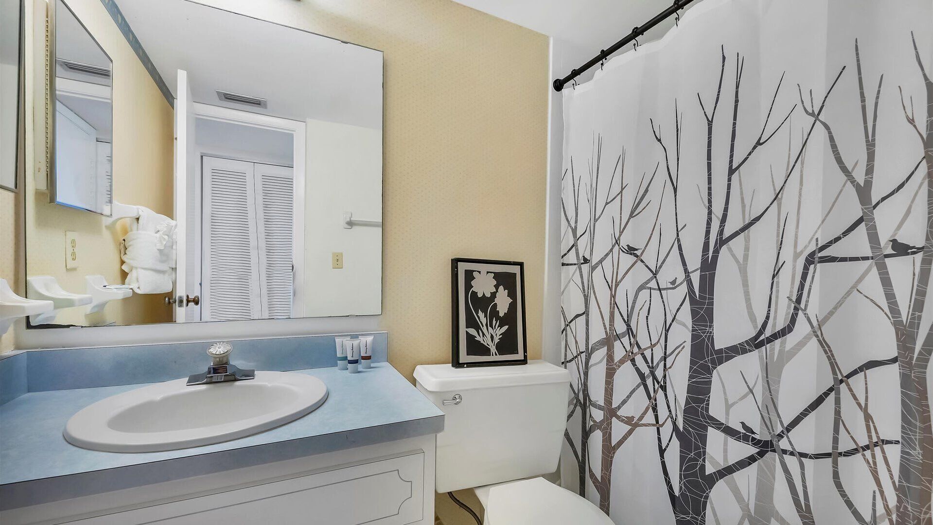 A bathroom with a sink, mirror, toilet, and shower curtain with a tree design.