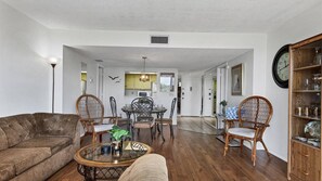 A spacious living room with a brown sofa, a glass coffee table, wicker chairs, and a dining area with a black table and chairs.