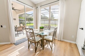 A dining area with a glass table and wooden chairs, large windows with blinds, and a view of a backyard.