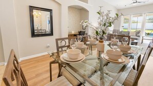 A dining area with a glass table set for six, surrounded by chairs. There is a large mirror on the wall, a bookshelf, and a window with a view of the outdoors. - Windsor Palms Resort Vacation Home WP2209 (Kissimmee)