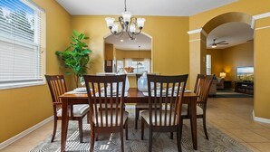 A dining area with a table set for six, surrounded by wooden chairs. A chandelier hangs from the ceiling, and a potted plant is visible to the left. - Windsor Hills Resort Vacation Home  TS7718 (Kissimmee)