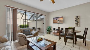 A spacious living room with a sliding glass door leading to a balcony, a comfortable sofa, a coffee table, a dining area with chairs, and a flat-screen TV mounted on the wall. - Windsor Palms Resort Vacation Townhome 3BR (Kissimmee)