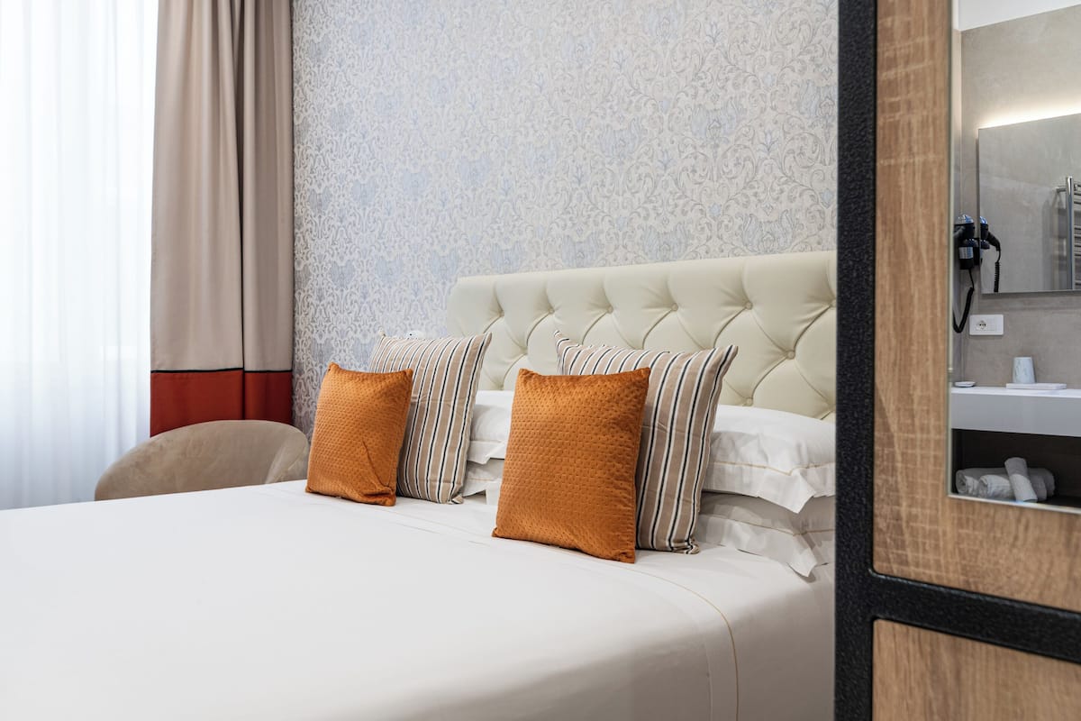A hotel room with a tufted headboard, a bed with striped and solid pillows, a bedside table, and a mirror.