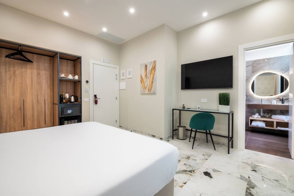 A hotel room with a bed, a desk, a TV, a mirror, and a bathroom visible through an open door.