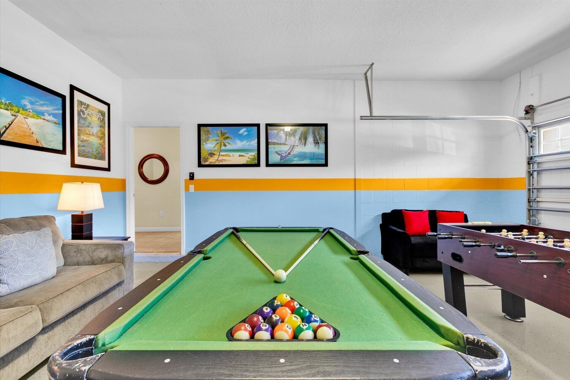 A pool table with billiard balls, a foosball table, and a sofa in a room with a blue and yellow stripe wall.