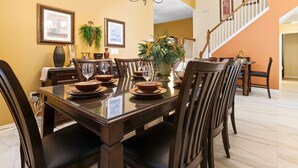 Une salle à manger avec une grande table dressée pour six personnes, entourée de chaises en bois. Un escalier mène à un autre niveau, et les murs sont peints dans des tons chauds.