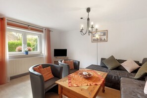 Ein Wohnzimmer mit einer Couch, Sesseln, einem Tisch mit einer Schüssel Essen, einem Fernseher und einem Fenster mit Vorhängen.