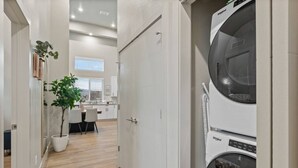 Une buanderie équipée d’une machine à laver et d’un sèche-linge superposés, avec un plancher en bois et une porte donnant sur un coin cuisine comprenant une table et des chaises.