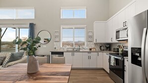 Une cuisine moderne avec des meubles blancs, des appareils en acier inoxydable et un coin repas comprenant une table en bois et un vase rempli de plantes vertes.