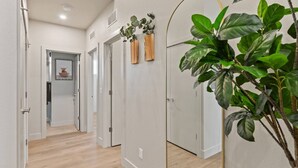 Un couloir doté d’un plancher en bois, de murs blancs et de portes donnant sur d’autres pièces. On y trouve des plantes en pot et un miroir accroché au mur.