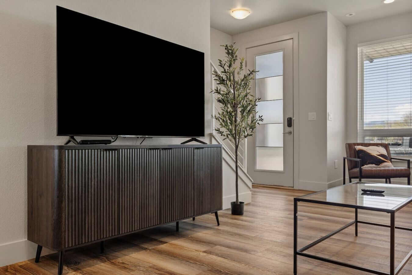 Een moderne woonkamer met een grote flatscreen televisie bevestigd aan een donker houten kast, een glazen salontafel, een potplant en een leren fauteuil.