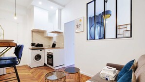 Un espace de vie compact comprenant un coin cuisine, un lave-linge, un coin repas avec une table et des chaises, et un canapé avec une petite table d’appoint.
