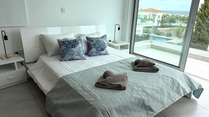 Ein modernes Schlafzimmer mit einem großen Bett, zwei Kissen, einem Nachttisch und Meerblick durch ein großes Fenster.