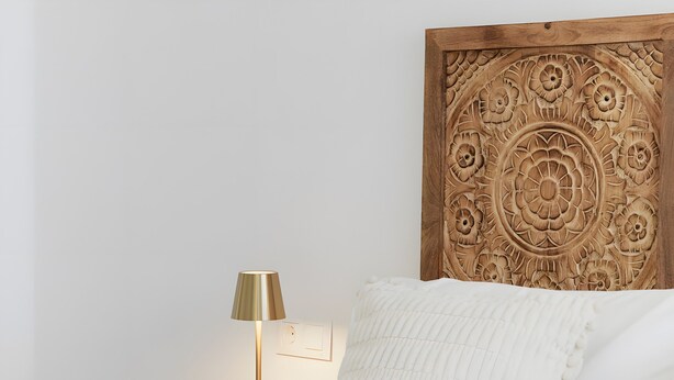 A neatly made bed with white linens, a wooden headboard with intricate carvings, a bedside lamp, and a small wooden nightstand.