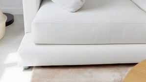 A white sofa with a single pillow, a framed picture on the wall, and a small round table with a vase.
