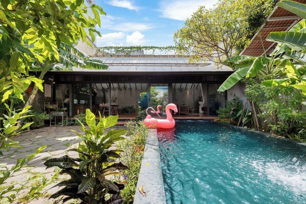 Ein Swimmingpool mit einem Flamingo-Schwimmding, umgeben von üppiger Vegetation und einer überdachten Sitzgelegenheit im Freien.
