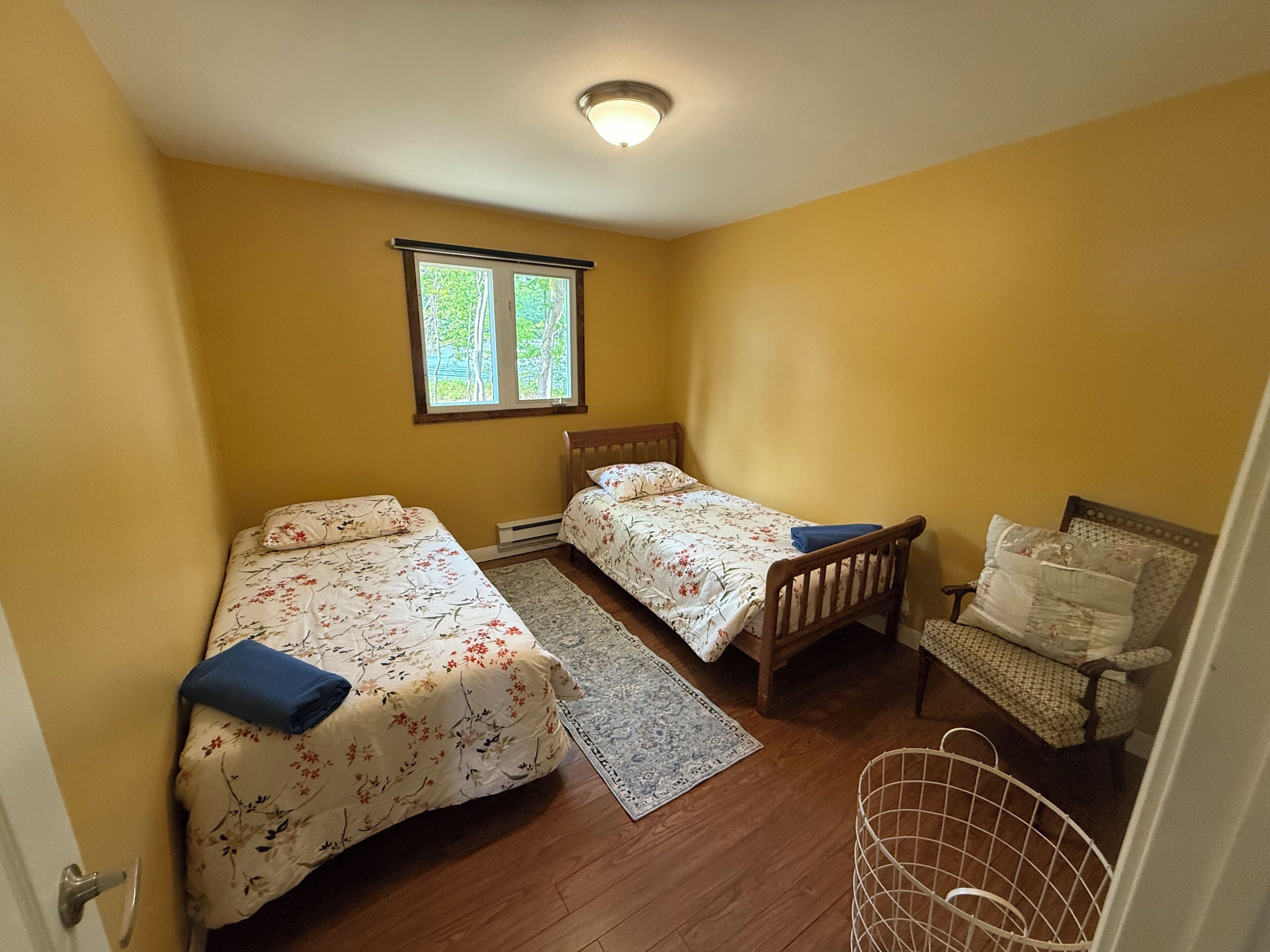 A small bedroom with two single beds, a wooden chair, a small table, and a window with a view of trees.