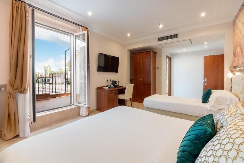 A hotel room with a large bed, a desk, a chair, a TV, and a balcony with curtains.