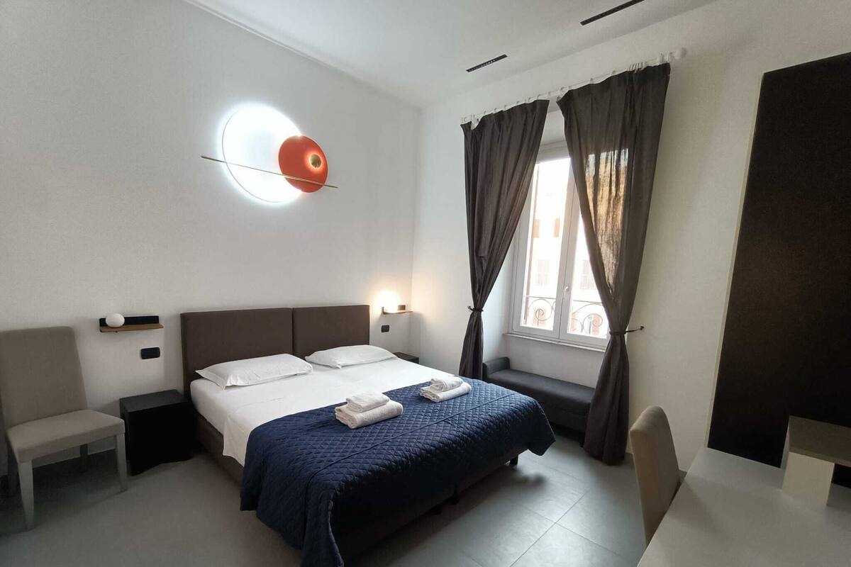 A hotel room with a bed, bedside tables, a wall-mounted light, a window with curtains, and a view of the street.