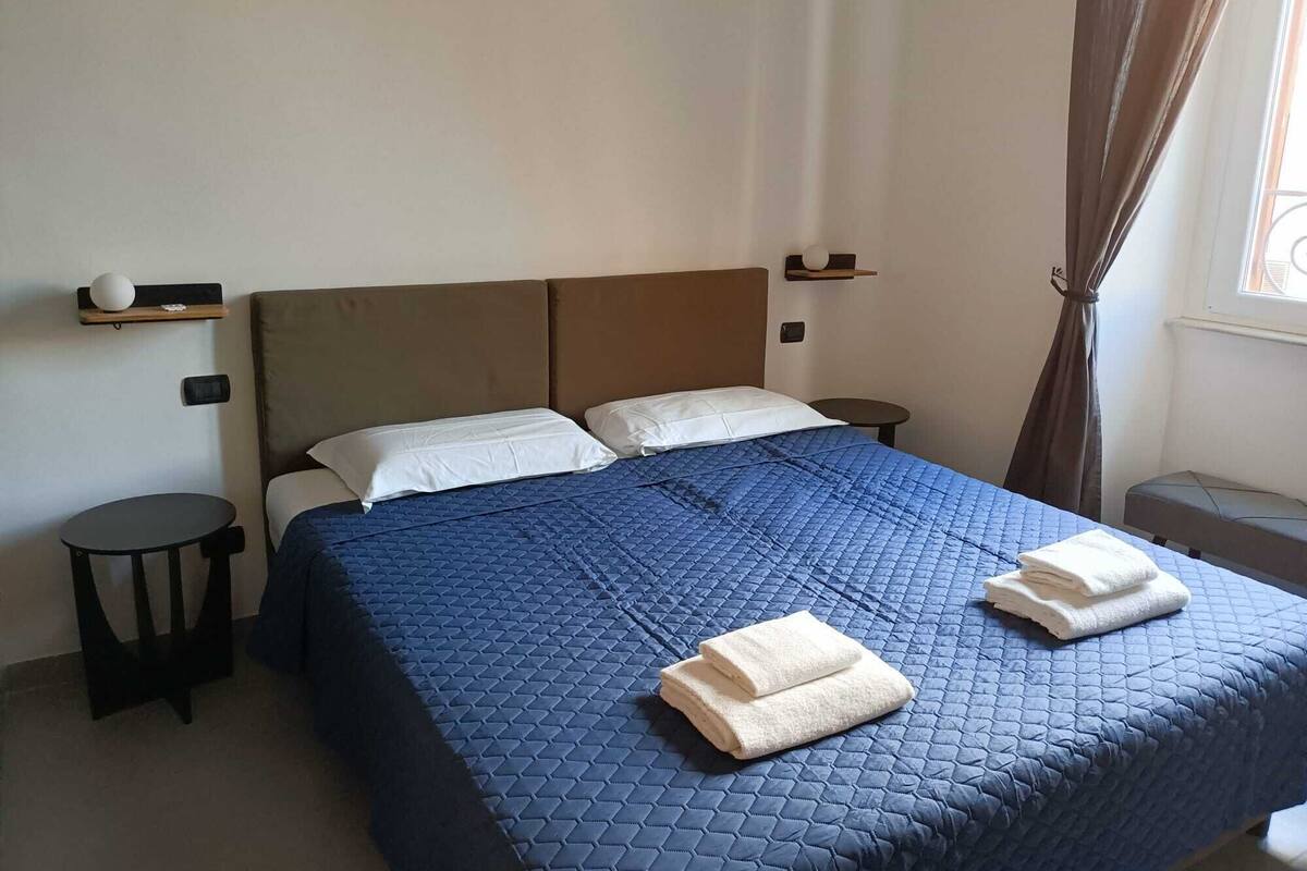 A neatly made bed with a blue quilt, two pillows, and two folded towels. There is a small bedside table with a lamp, a wall-mounted shelf with a decorative item, and a window with curtains.
