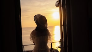 A person wearing a hat, standing by a window with a view of the ocean.