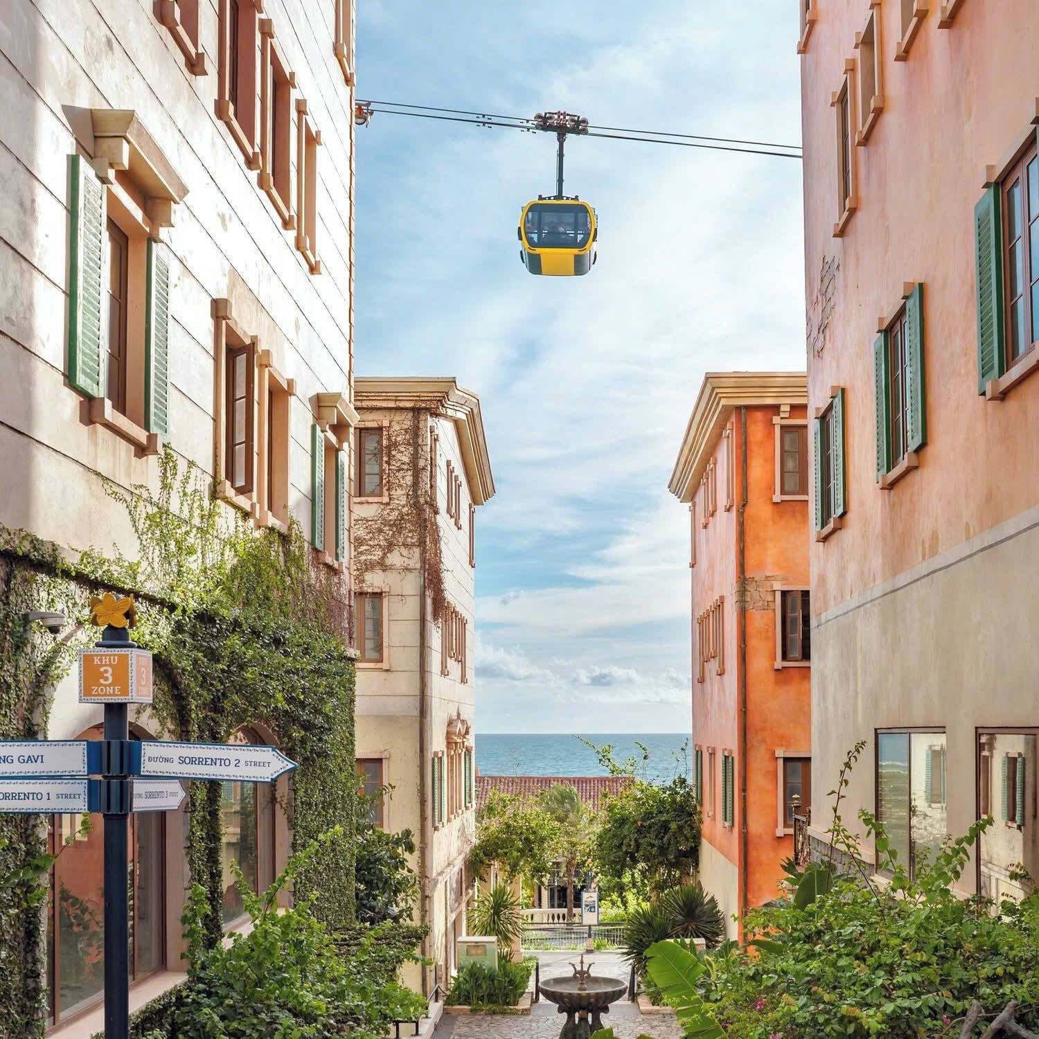 Eine Seilbahn fährt zwischen Gebäuden, ein Schild weist 'Khu 3 Zone' aus, und Straßenschilder zeigen 'Duong Sorrent 2 Straße' und 'Duong Sorrent 3 Straße'.