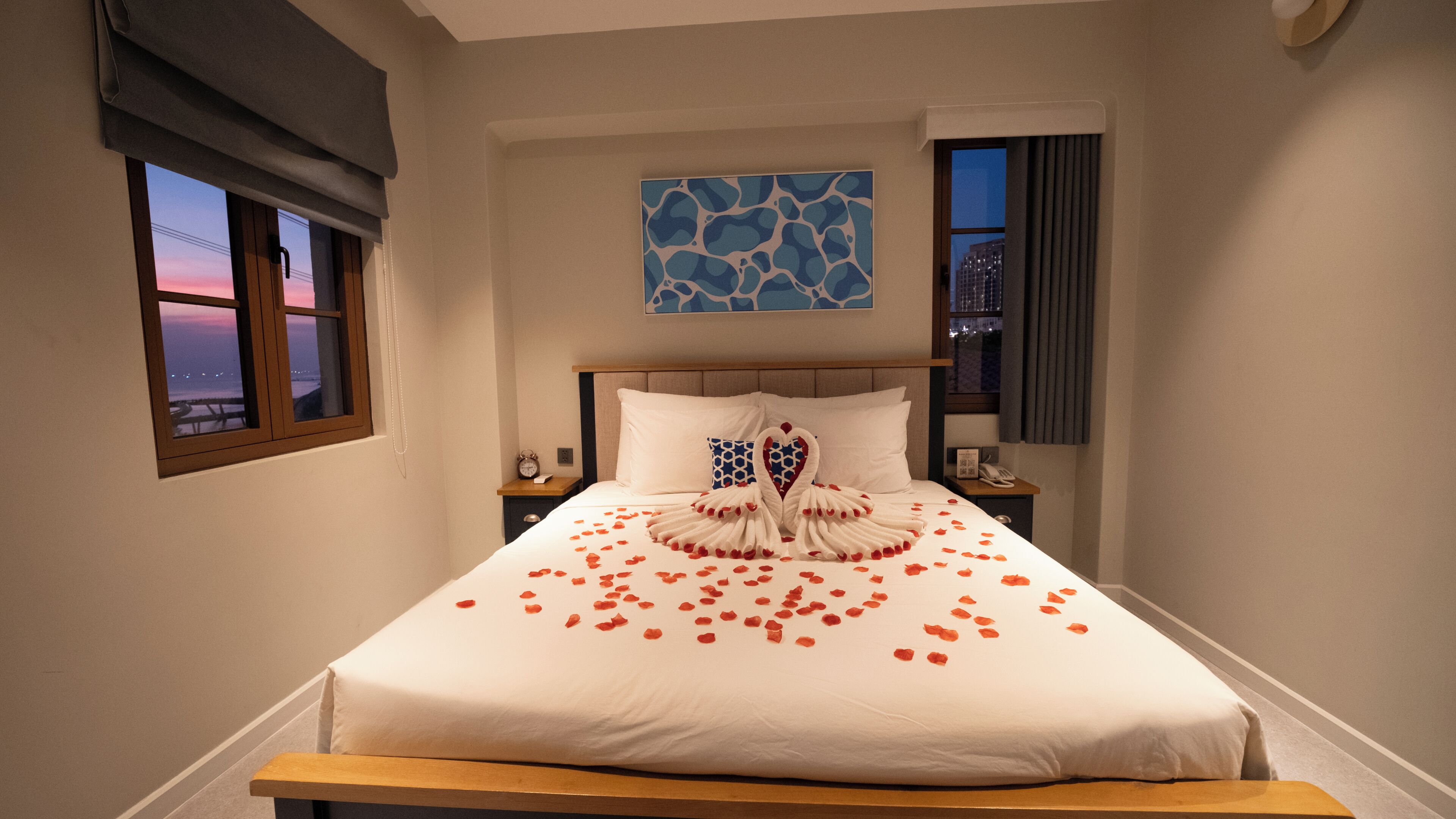 A hotel room with a bed, a nightstand, a clock, a phone, a window with a view, and a painting on the wall.