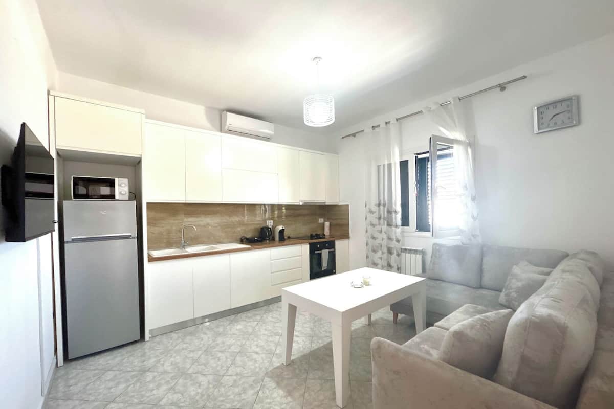 Une cuisine compacte avec des meubles blancs, un four à micro-ondes encastré et une cuisinière à gaz. On y trouve une table blanche et un canapé beige dans l’espace de vie.