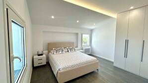 A modern bedroom with a large bed, a nightstand, a wardrobe, and a window with a view of greenery. - Ishëm Greenview Apartments (Ishëm)