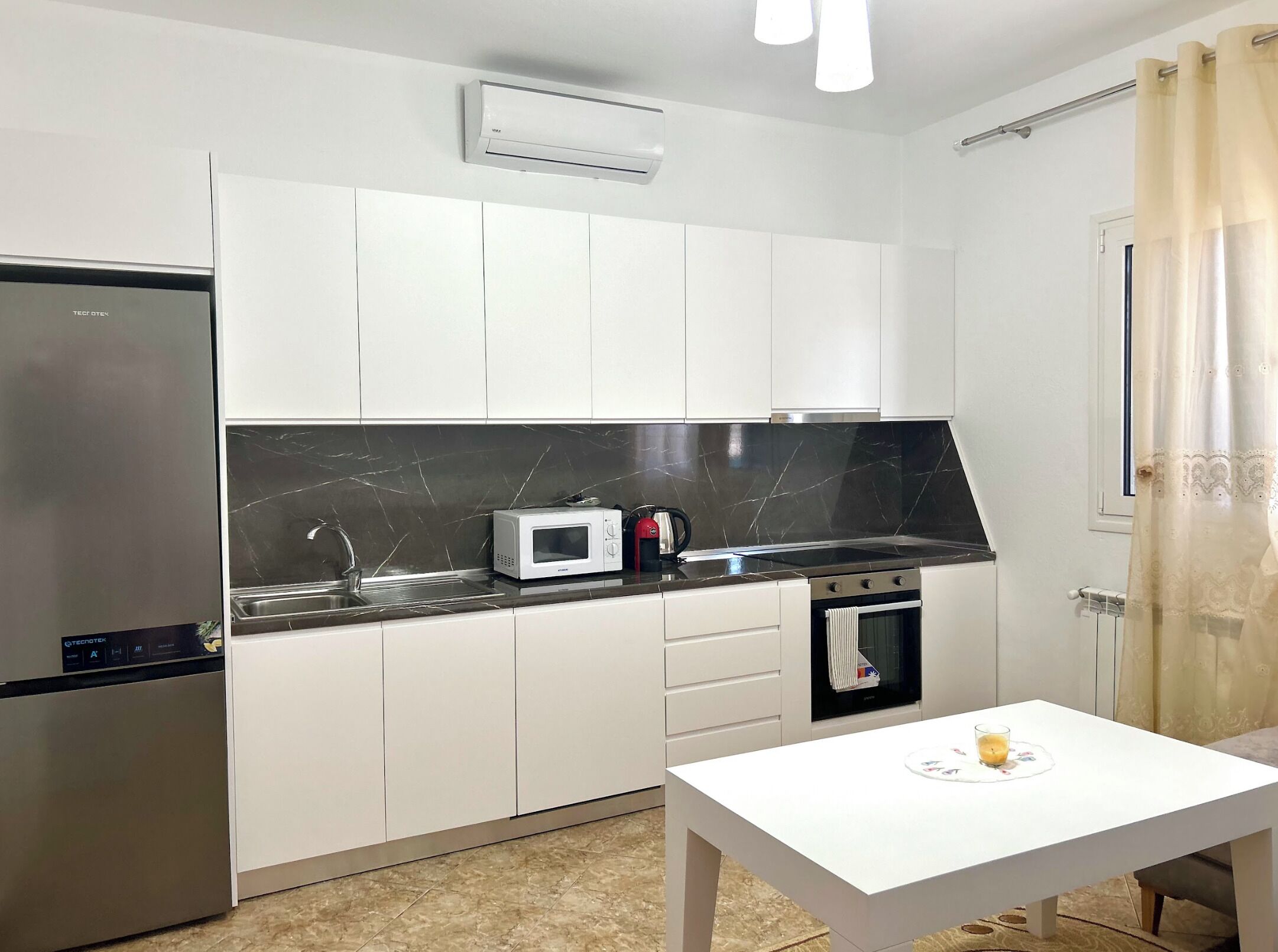 Uma cozinha compacta com armários brancos, bancada preta, micro-ondas, chaleira, fogão e uma pequena mesa com uma vela.