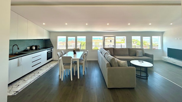 A spacious living area with a dining table, sofa, and coffee table, large windows with a view, and a modern kitchen. - Ishëm Greenview Apartments (Ishëm)